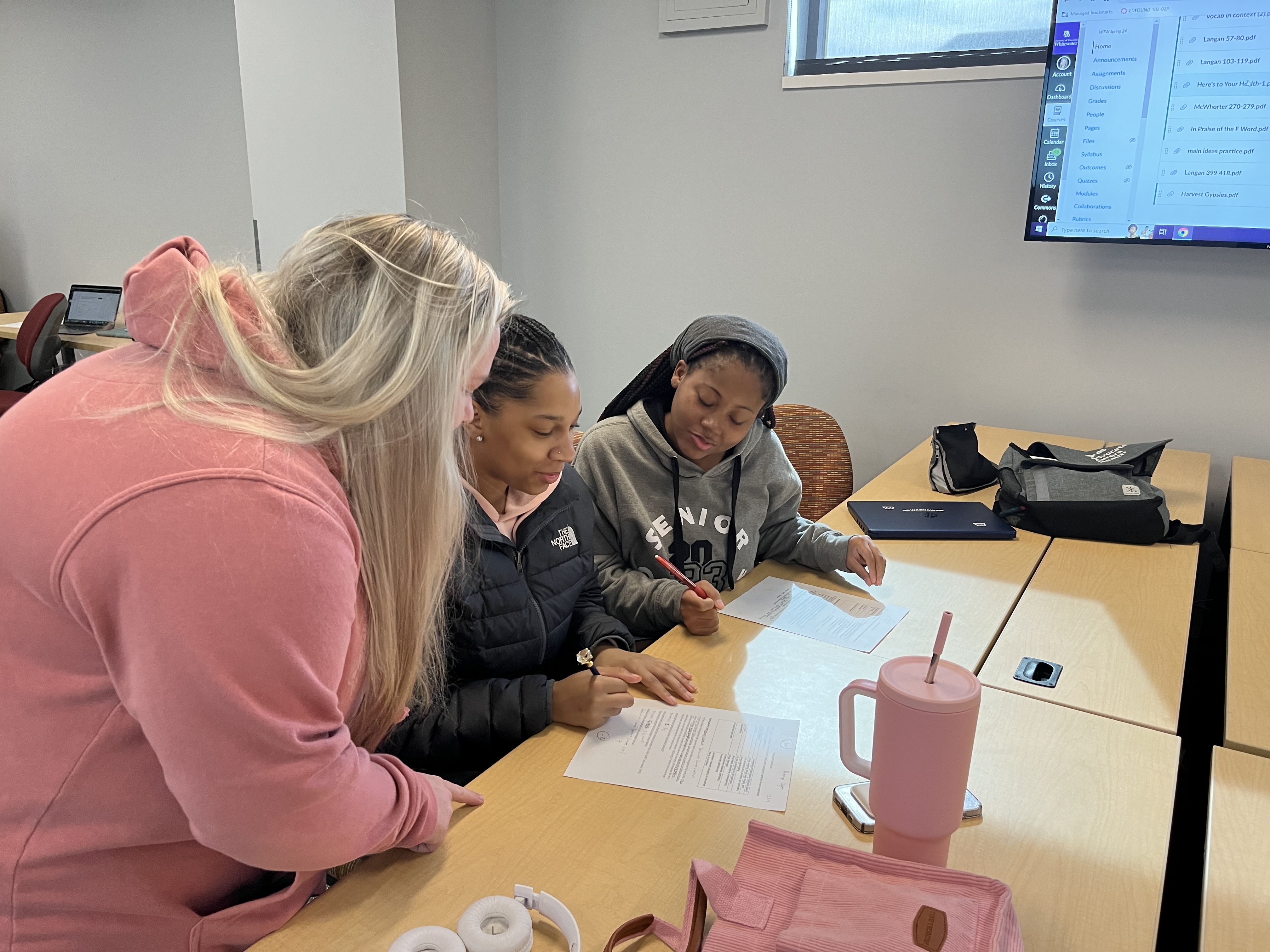 Students reviewing papers at a table with a TV in the background and a pink mug on the table.