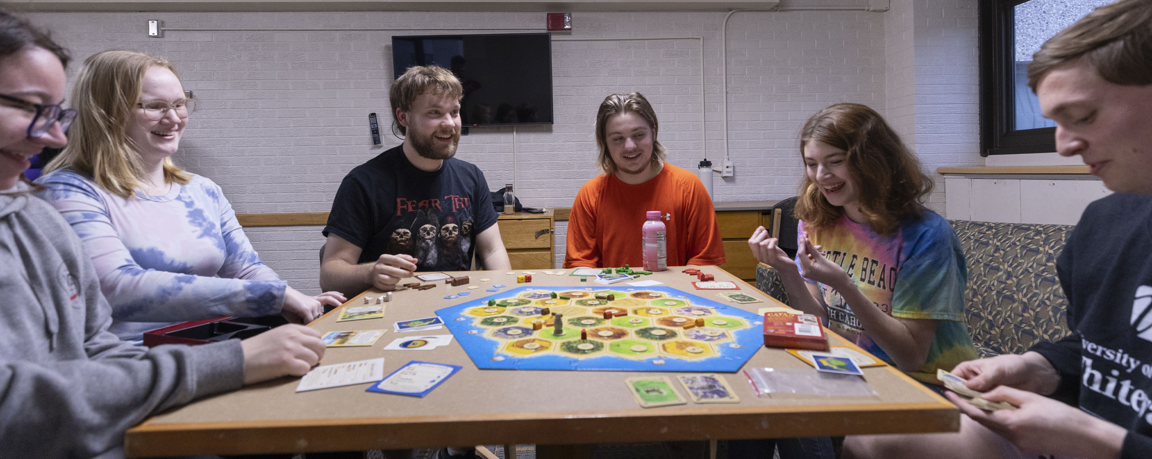 Six students laugh together as they sit at a table with a game board on it.