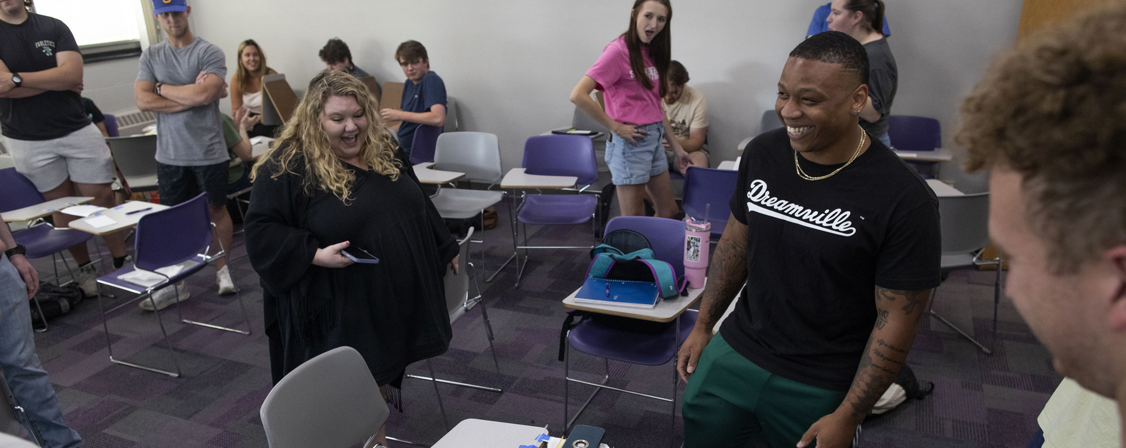 Students in a classroom cheer as one student smiles, standing amongst desks and chairs.