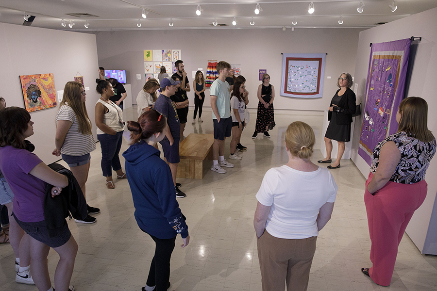 A group of students are gathered around a faculty member who is standing next to a large piece of artwork hanging on a gallery wall.