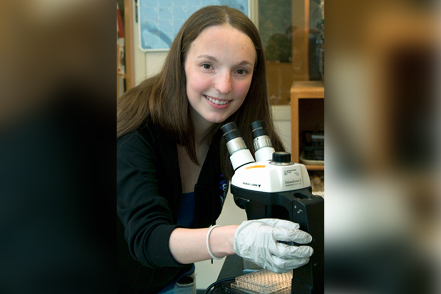 A woman looks up from a microscope and smiles at the camera.