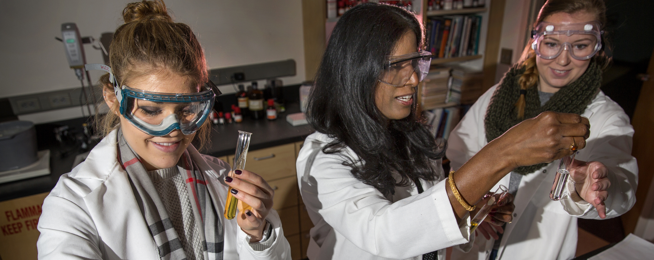 A faculty member and two students, all in lab coats, look at liquid vials.