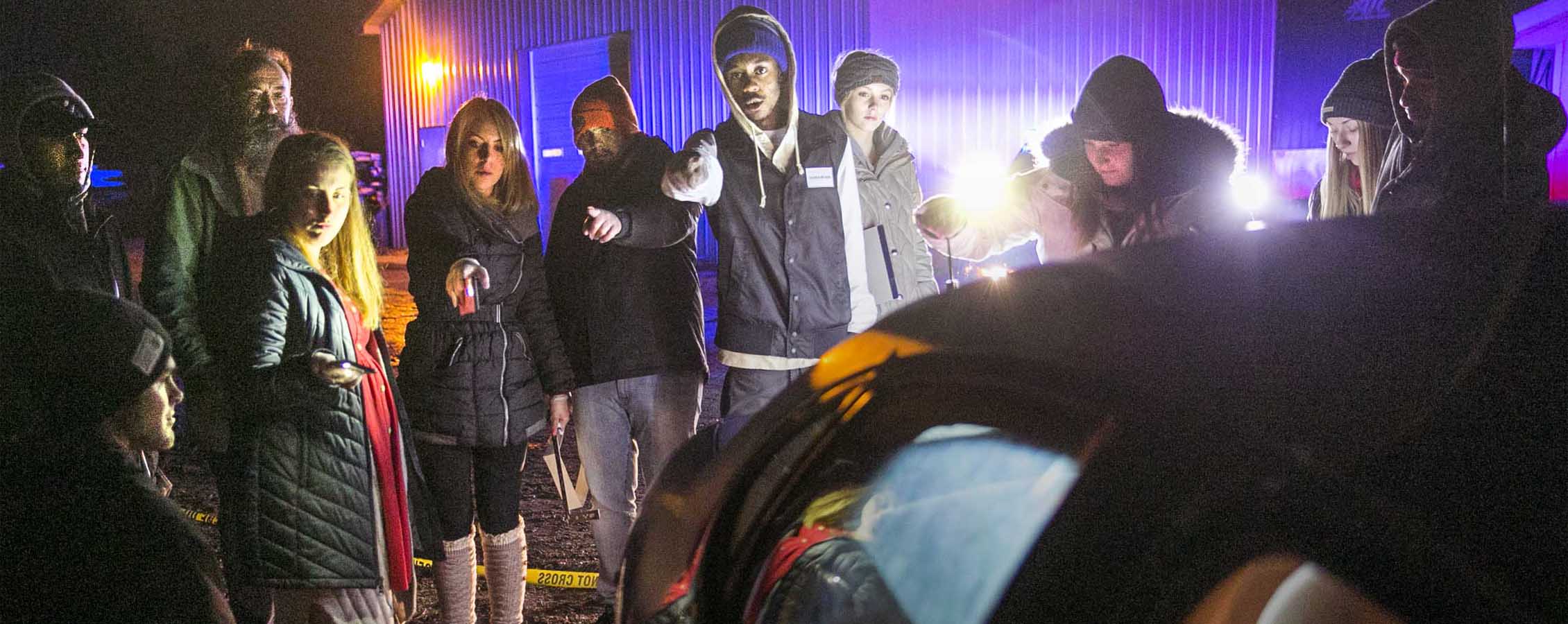 Students stand outside in the dark by a car during a crime scene simulation.