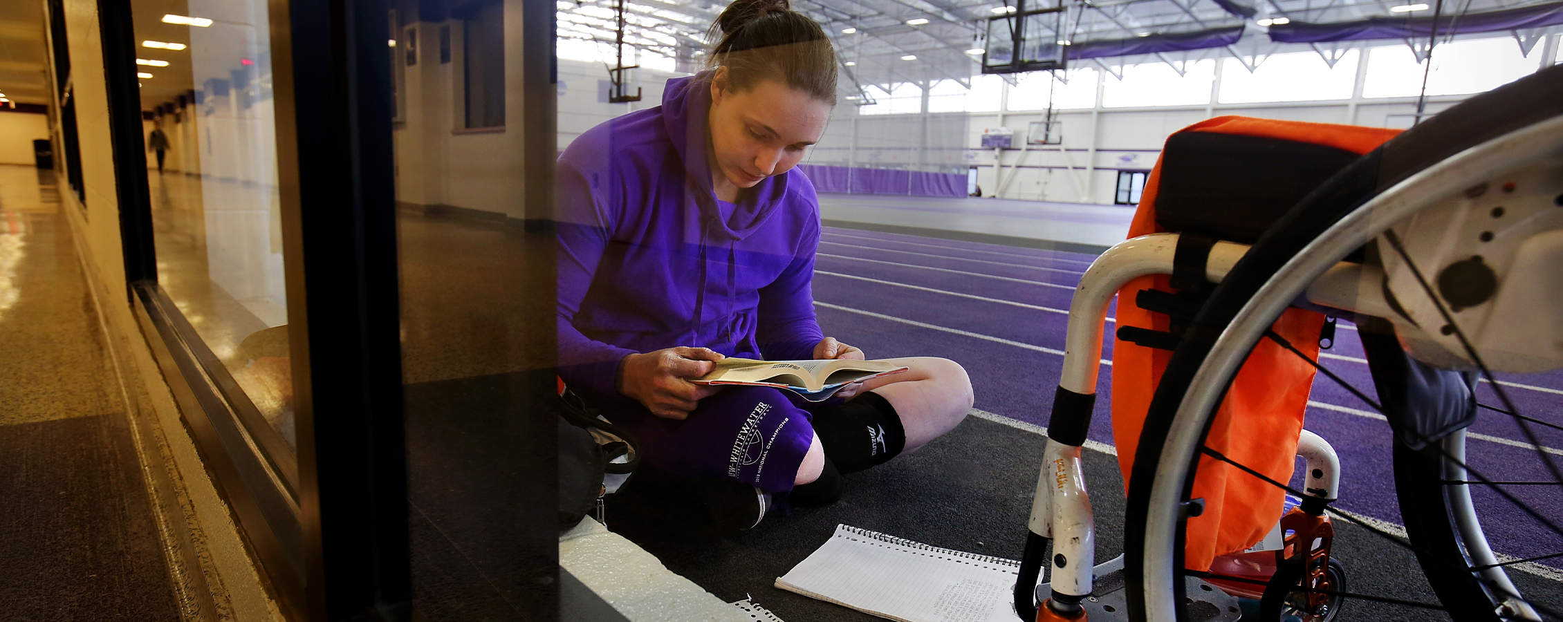 A person sits on the floor in front of their wheelchair as they read a book.