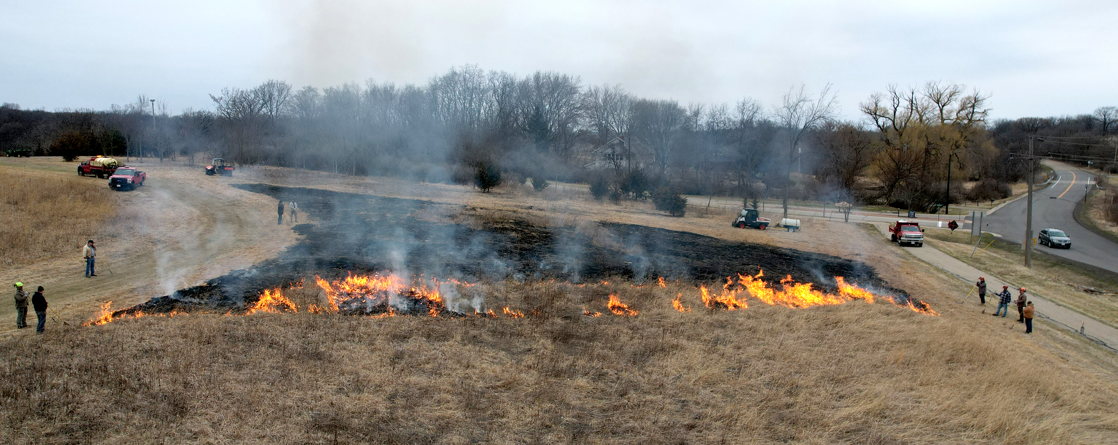 A drone photo shows people standing around a controlled fire in a prairie.