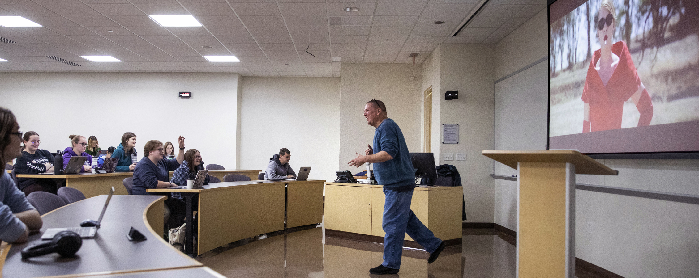 A faculty member speaks at the front of class while a film plays behind him.