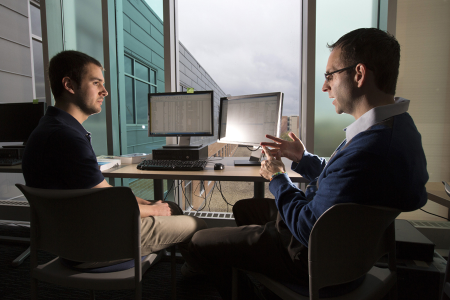 A student and a faculty member have a discussion at a table by two computers.