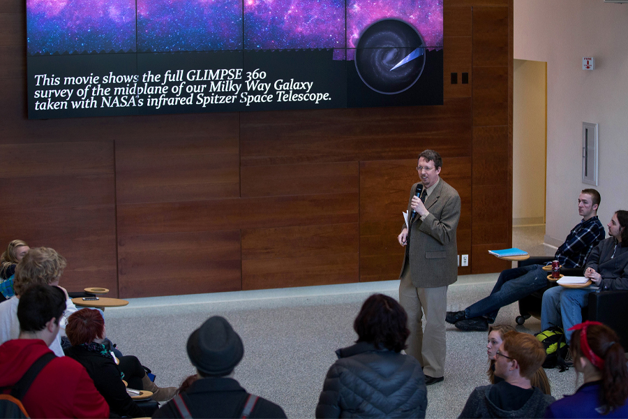 A faculty member speaks to students standing next to a presentation about the Spitzer Space Telescope.