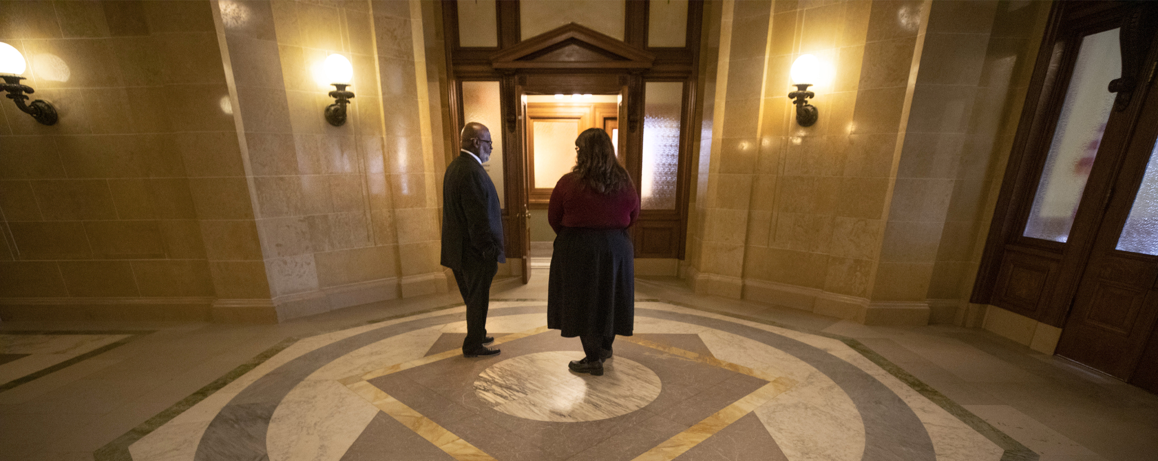 Two people talk in a room with dark woodwork and and American flag.