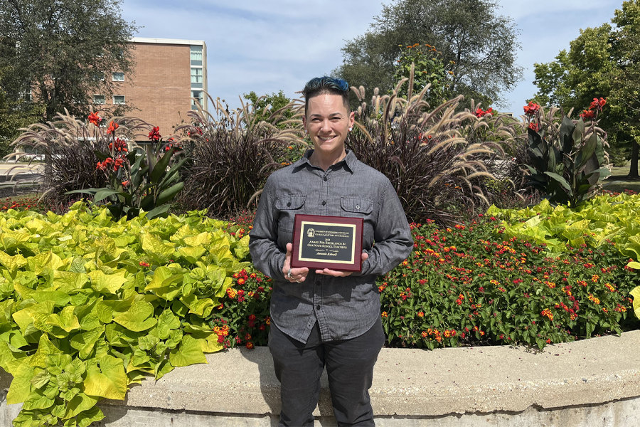 Amanda Kidwell stands outdoors amongst colorful plants and holds an award.