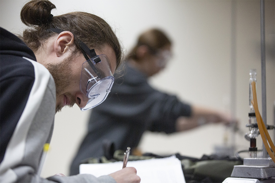 A student wearing safety goggles writes in a notebook.