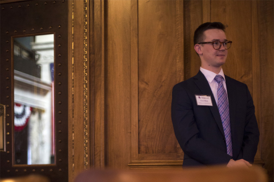 A student wearing a nametag stands against a beveled wood paneled wall.