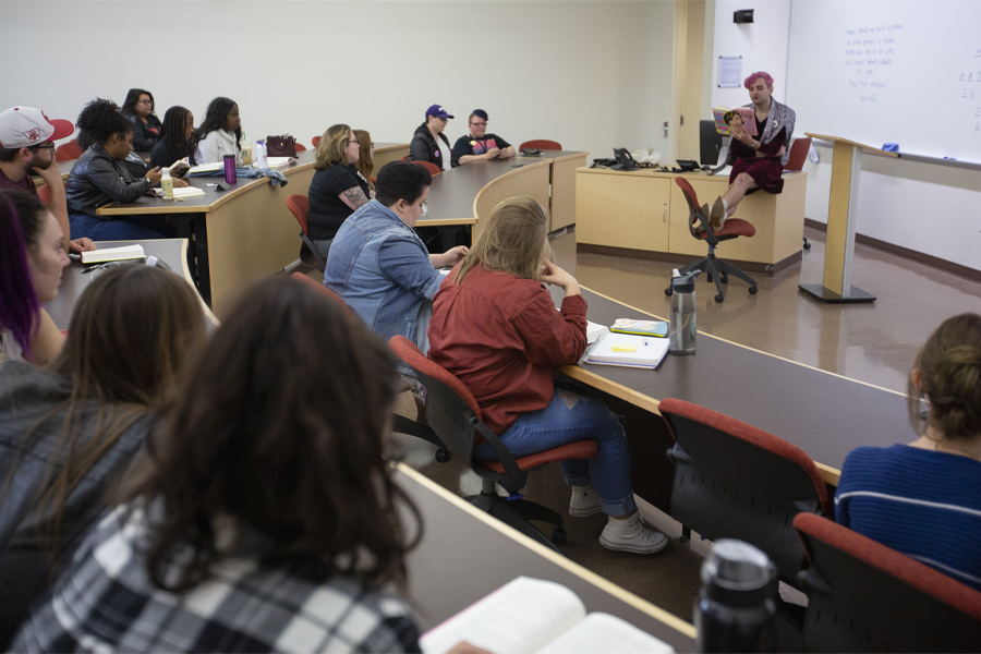 A person with pink hair reads from a book at the front of class.