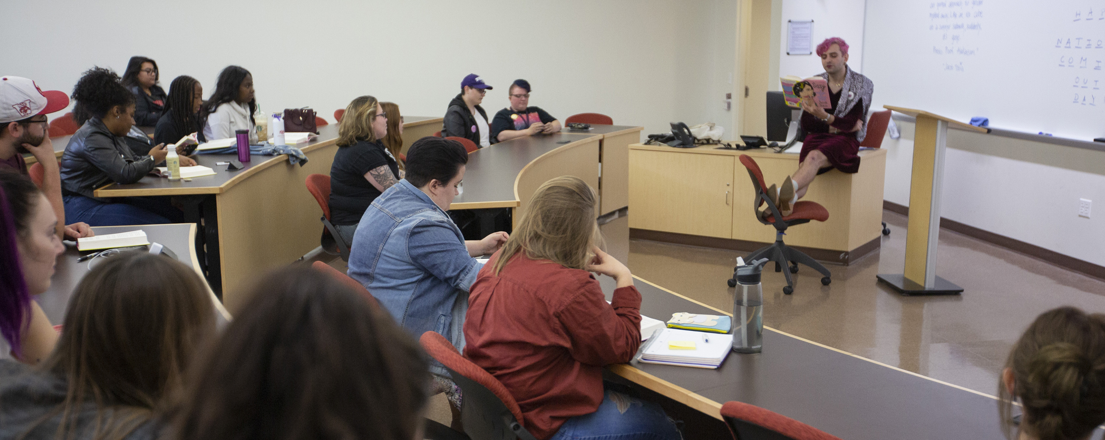 A person with pink hair reads from a book at the front of class.