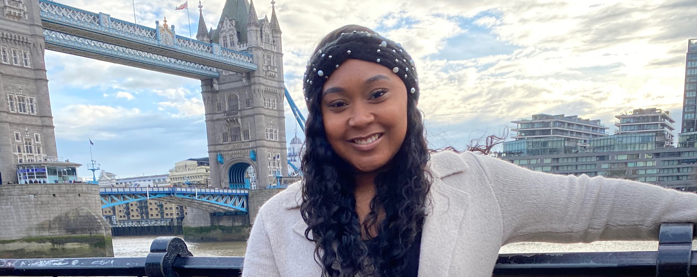 A student stands in front of a large bridge in a European city and smiles at the camera.