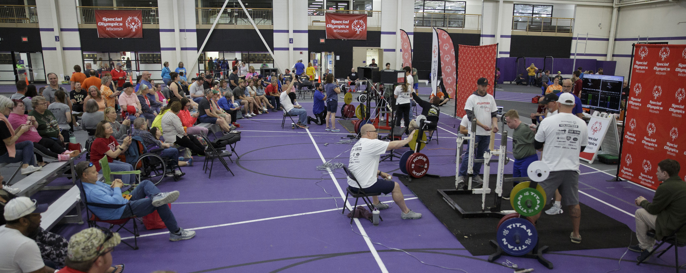 A crowd of people gathered in the fieldhouse watch special olympians do weight lifting competitions.