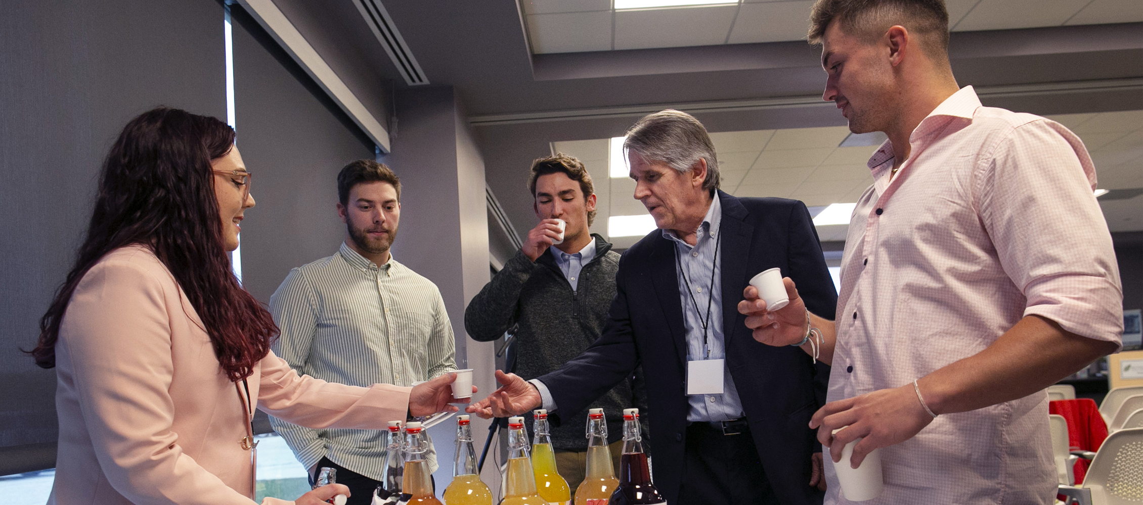 A student hands out cups of red and yellow kombucha to people gathered around a table.