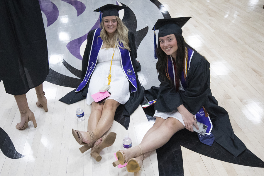 Two students wear academic regalia and sit on the gym floor by a large Warhawk head.