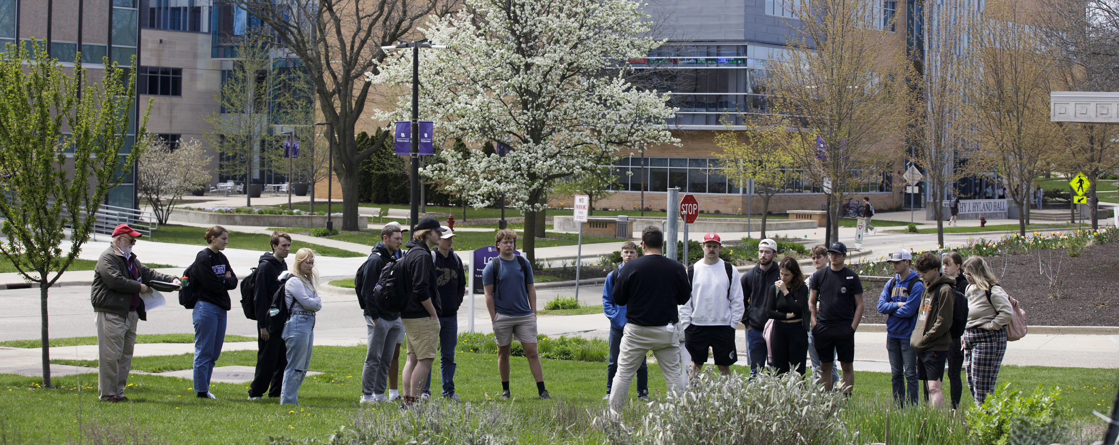 A class stands outdoors amongst buildings and trees with a faculty member speaking.