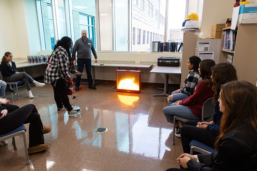 Students are gathered in a classroom with a simulated fireplace and one student is holding a fire extinguisher.