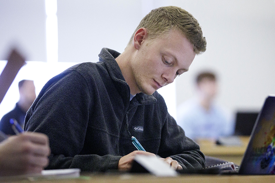 A student sits in front of a laptop and writes in a notebook.