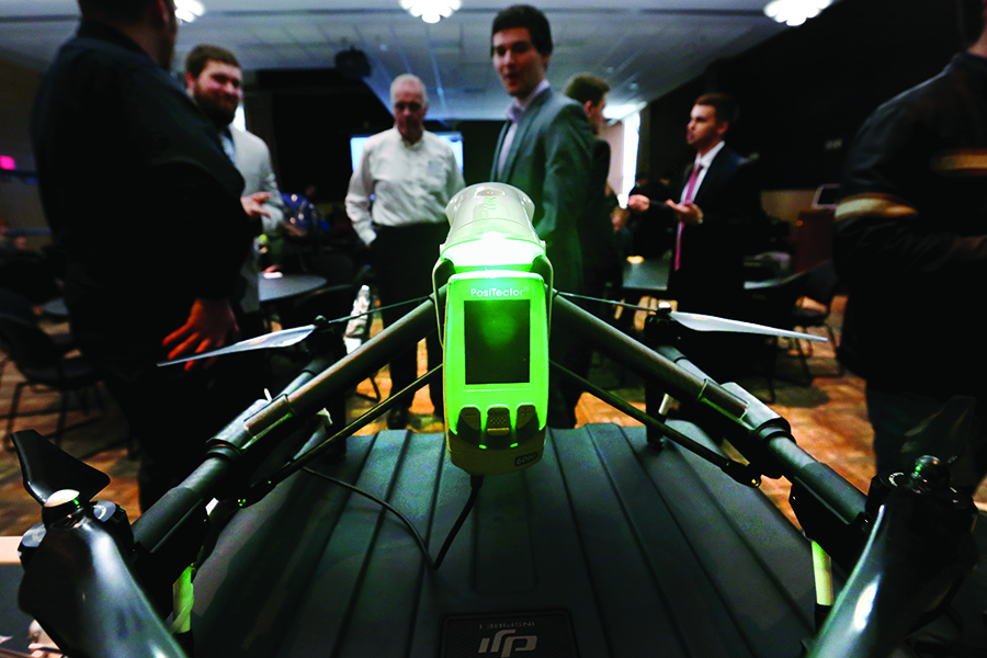 A green camera is mounted on a black drone with people in the background.