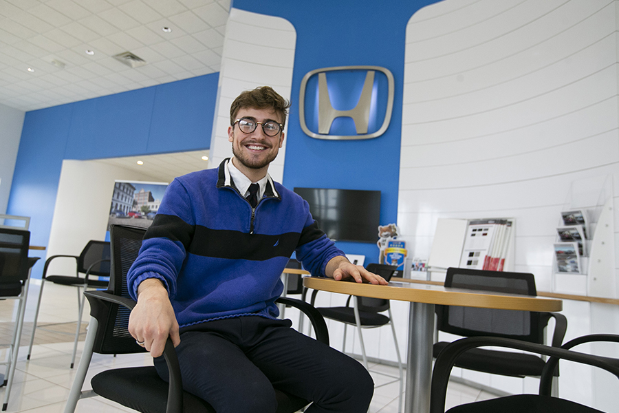 A student sits at a desk at a Honda dealership with a large Honda logo on the wall.