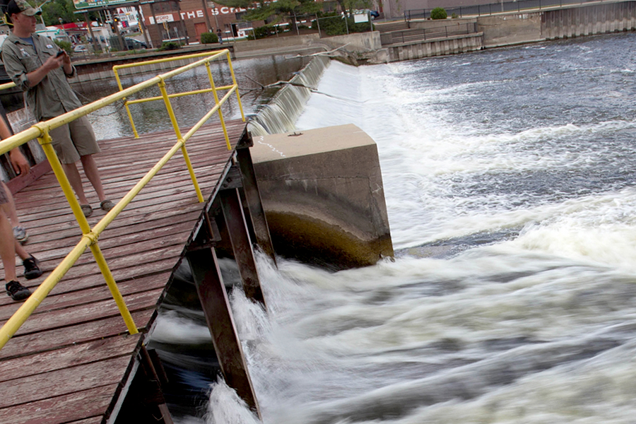 A student stands on a pier with a water testing device as water rushes underneath.