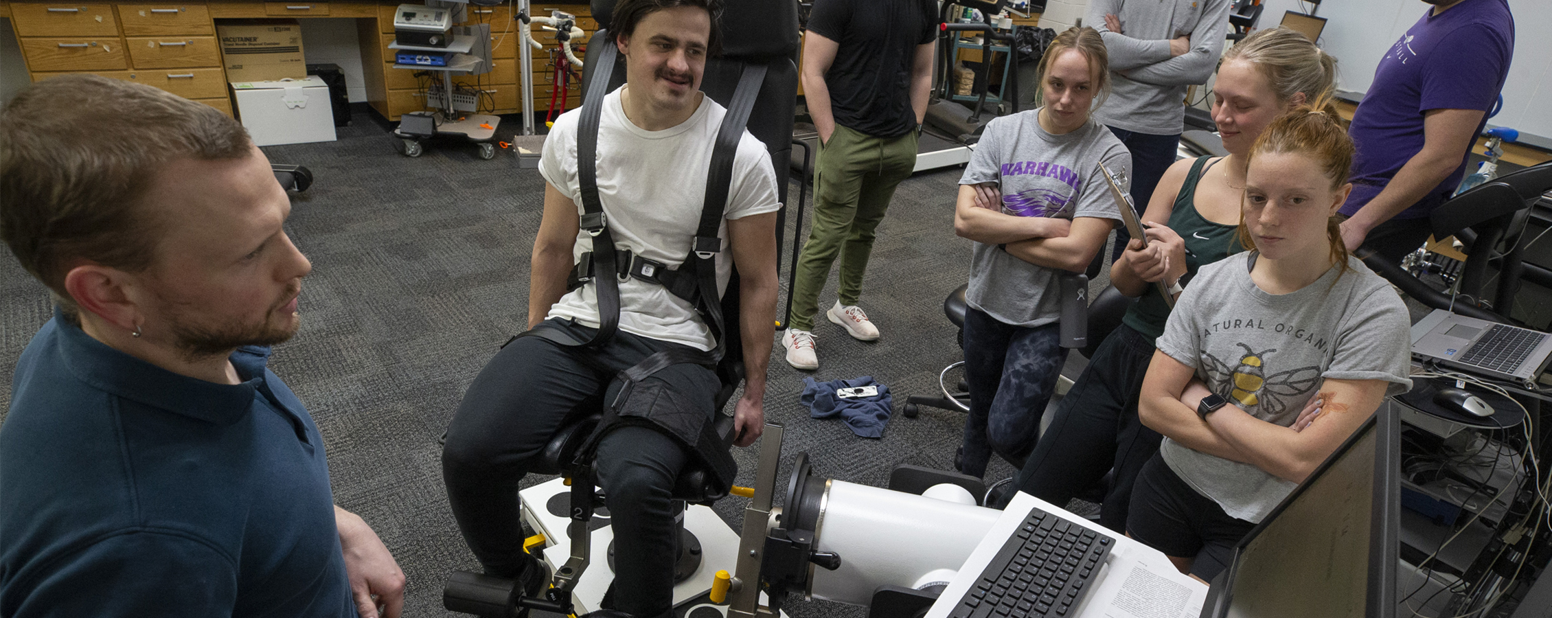 A faculty member and students gather around a student strapped in a chair to collect data during an exercise during class.