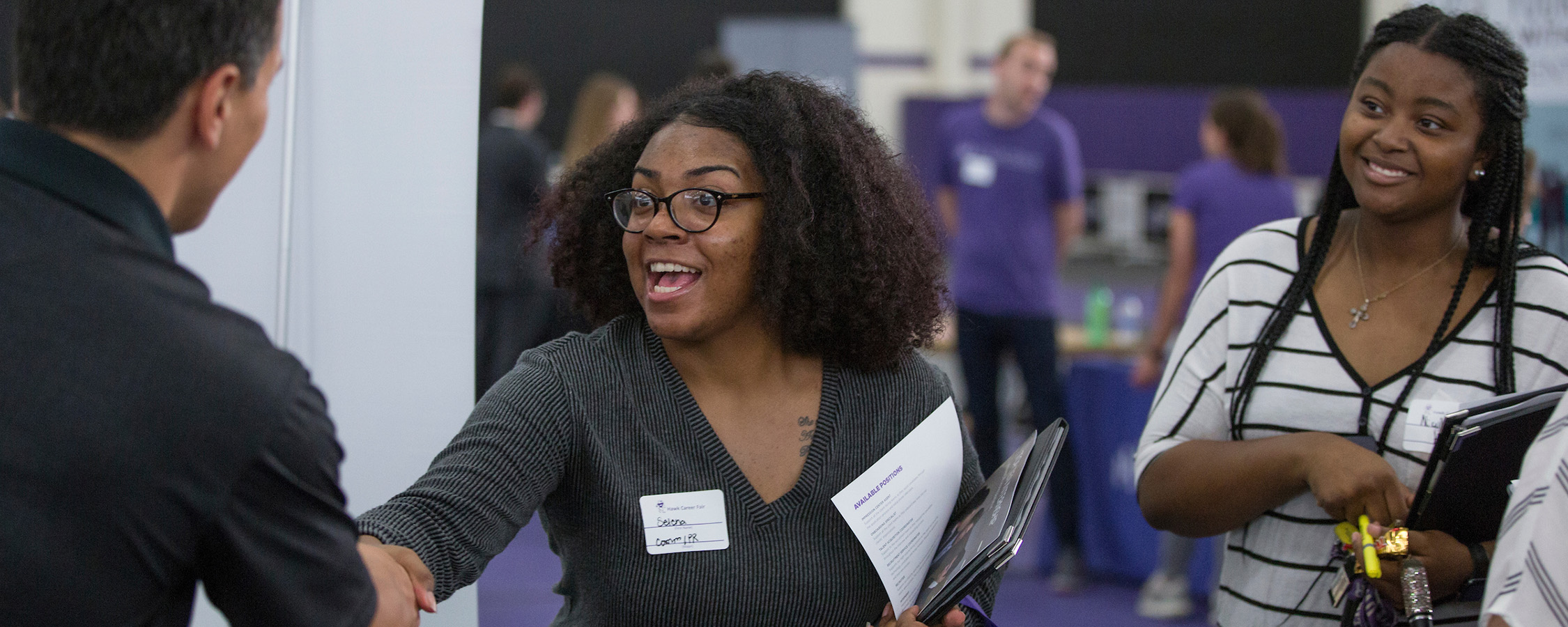 Two women at a career fair shaking hands with a man just out of the frame