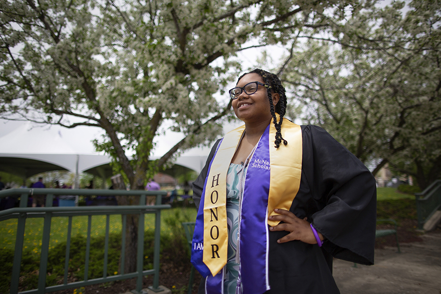 A student stands proudly in graduation attire with a yellow honors cord around her shoulders.