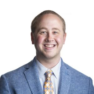 Brock Splittgerber wears a blue suit and gold tie while standing in front of a white wall.