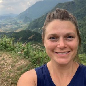 Dr. Jenna Cushing-Leubner smiles while wearing a blue tank-top while standing among green, tree-covered mountains.