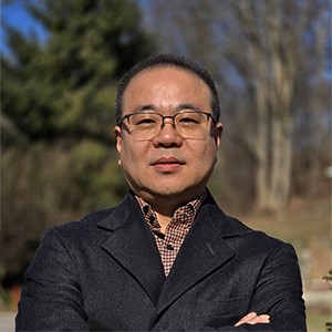 Headshot of Dan Kim outside wearing glasses, a checkered shirt, and a black suit coat