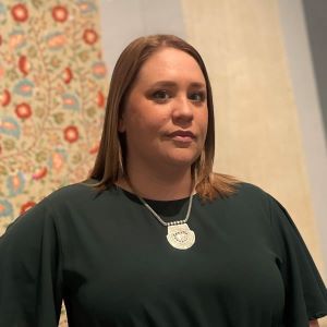 Ashley Dimmig stands in front of a wall with a flower print on it wearing a black shirt and a silver necklace.