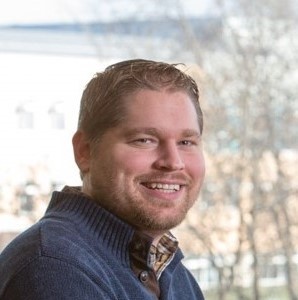 Jonah Ralstaon smiles while wearing a blue sweater with trees in the background.