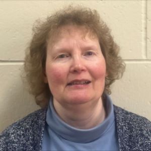 Elizabeth Jozwiak wearing a blue turtleneck and a blue sweater standing in front of a tan brick wall.