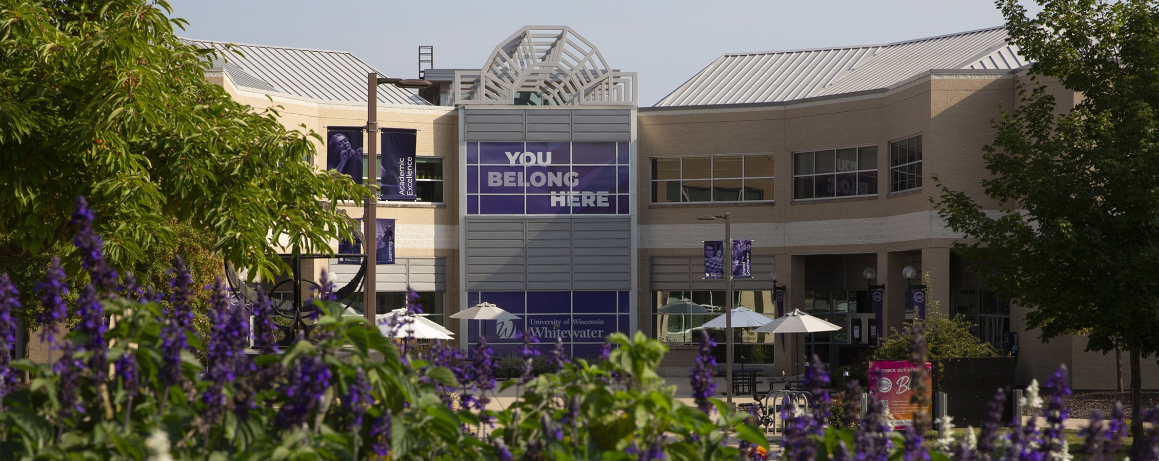 Exterior of the University Center with large green trees and flowers in the foreground.
