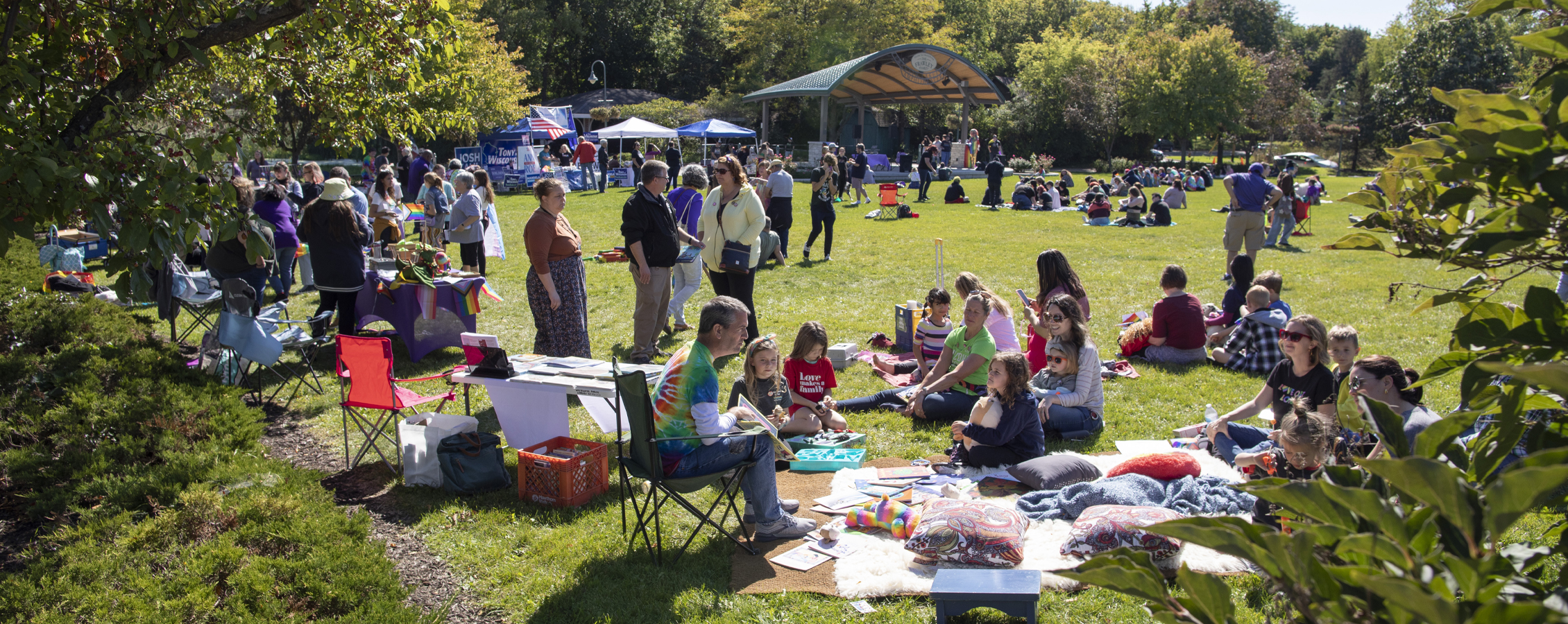 People gather in an outdoor green space surrounded by trees and pop up tents.