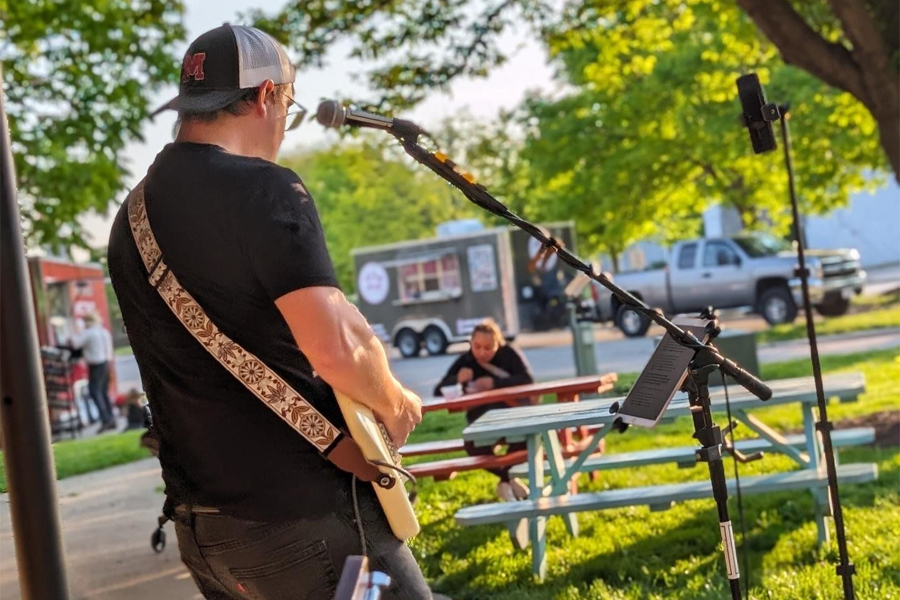 A person plays a guitar outdoors for a small crowd with a food truck in the background.