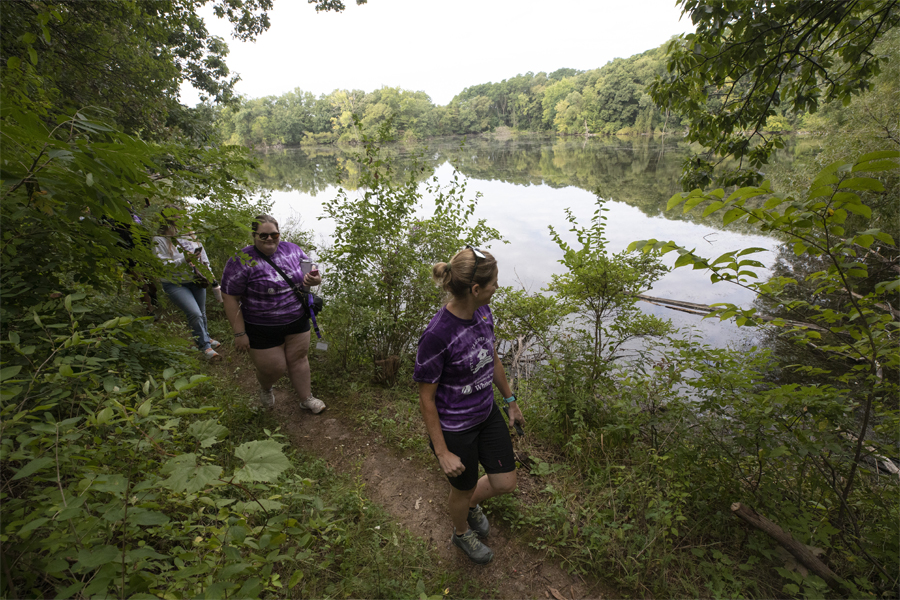 People walk along a trail through the woods and a clearing that shows a pond below.