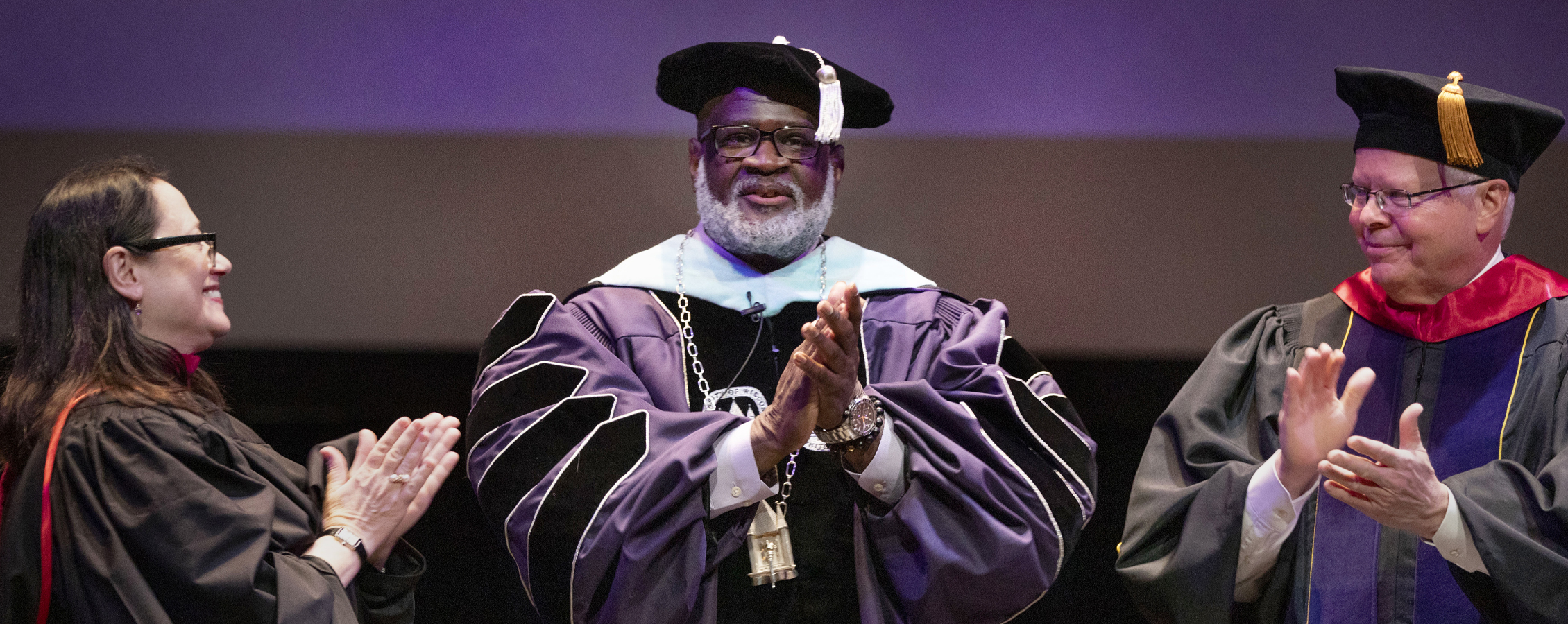 Chancellor King stands with two other people on stage, all dressed in academic regalia.