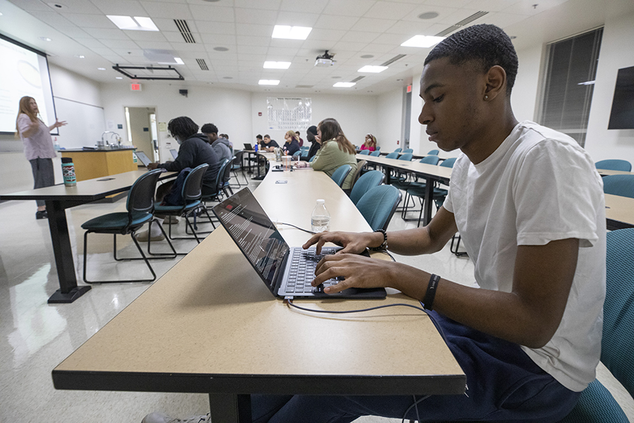 A student works on their laptop as a lecturer stands in the front of class.