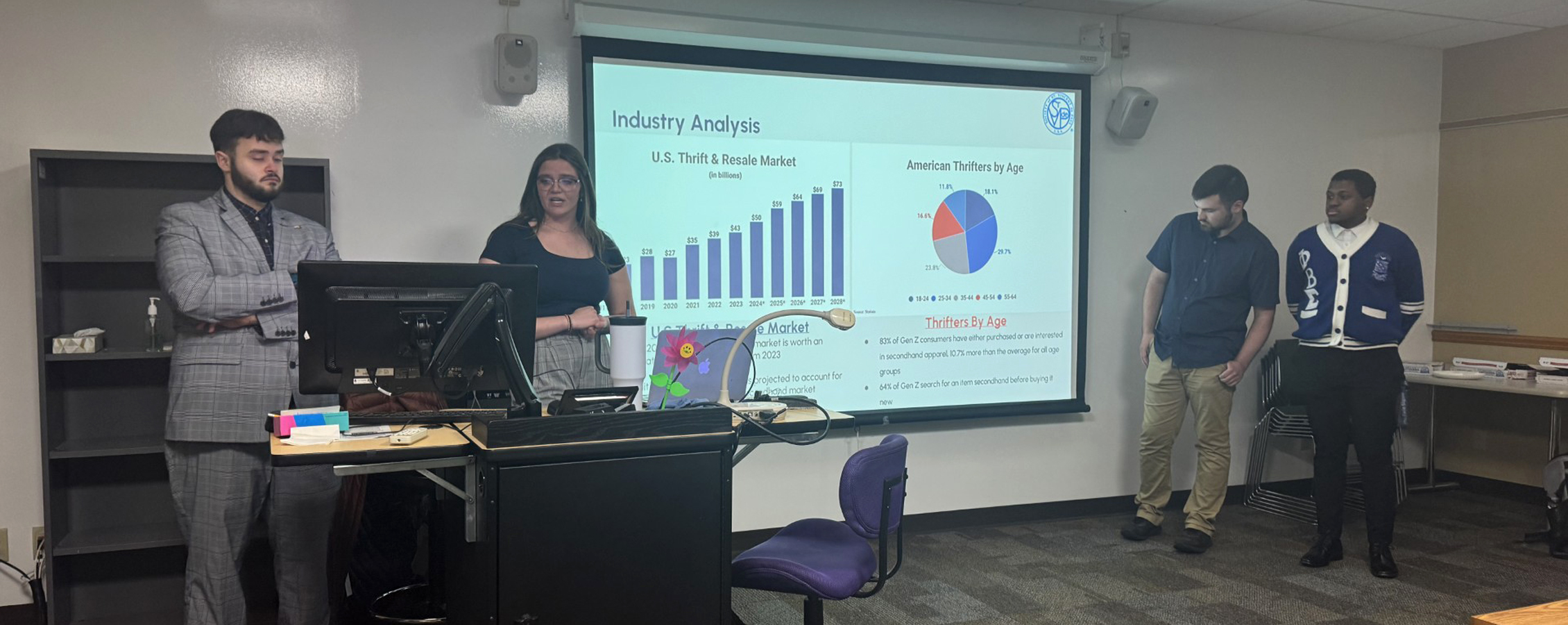 Four students stand in the front of a class and present work on a large screen.