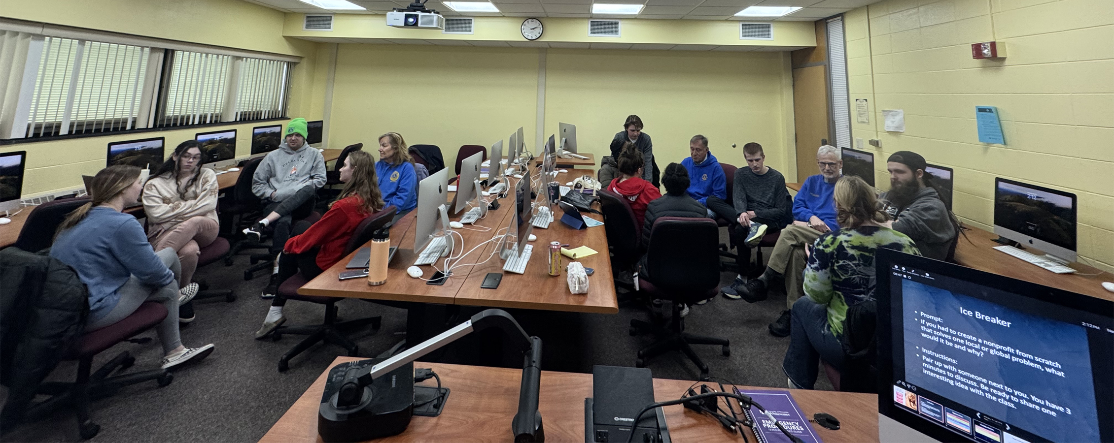 People gather in a classroom with computers on the tables.