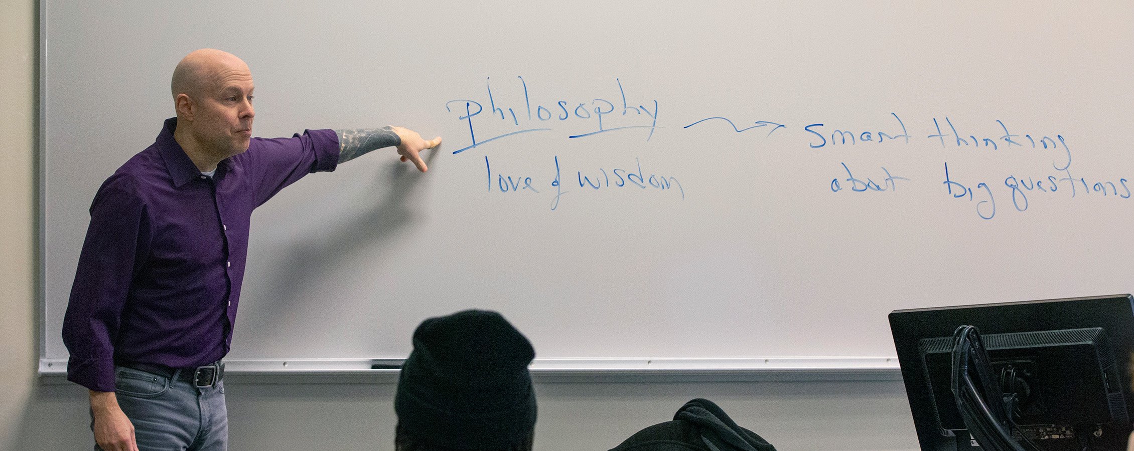 Chris Minor, professor of philosophy, pointing to the word Philosophy on a whiteboard.