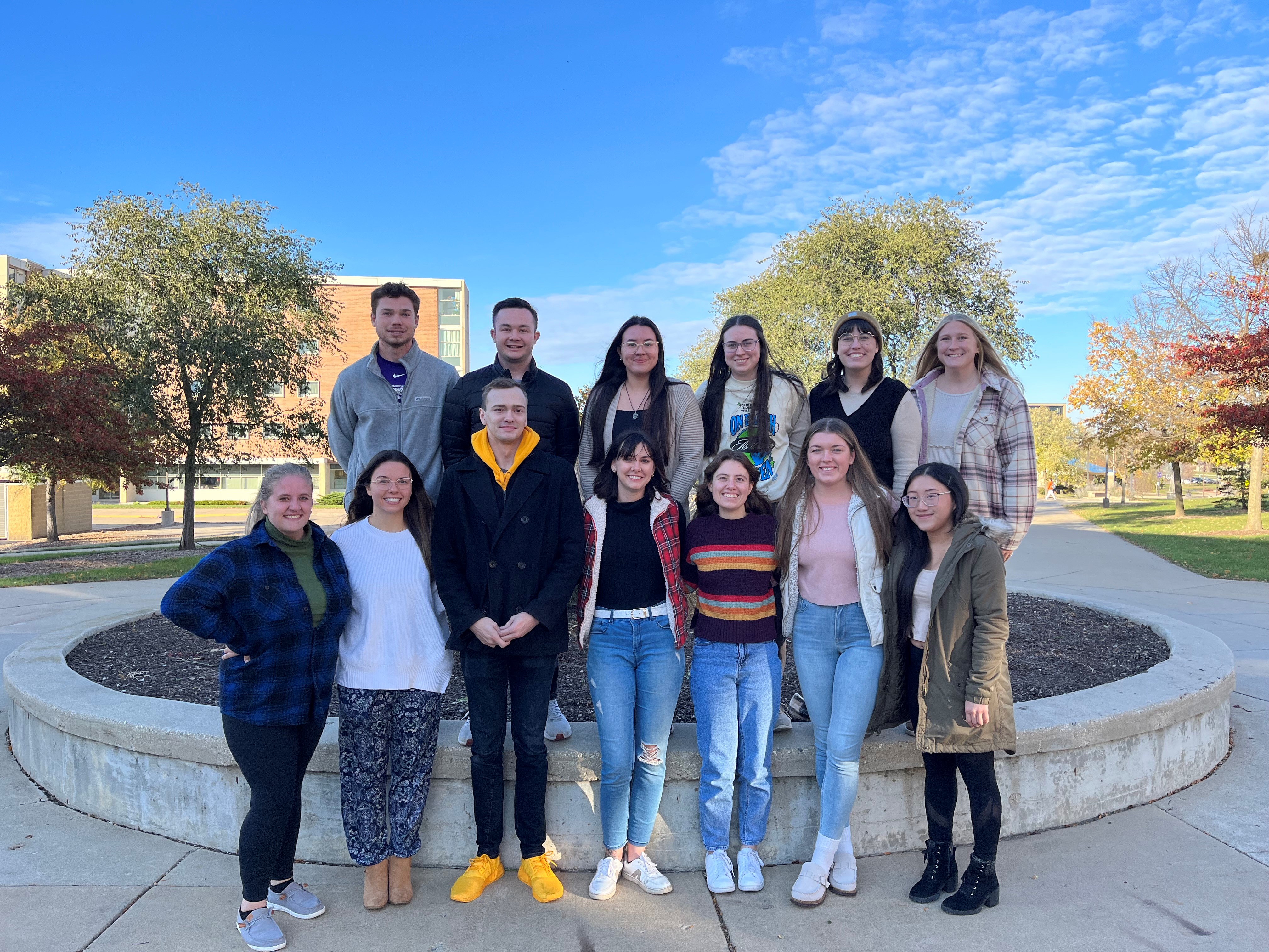 A group of people stand together in two rows outdoors on campus.