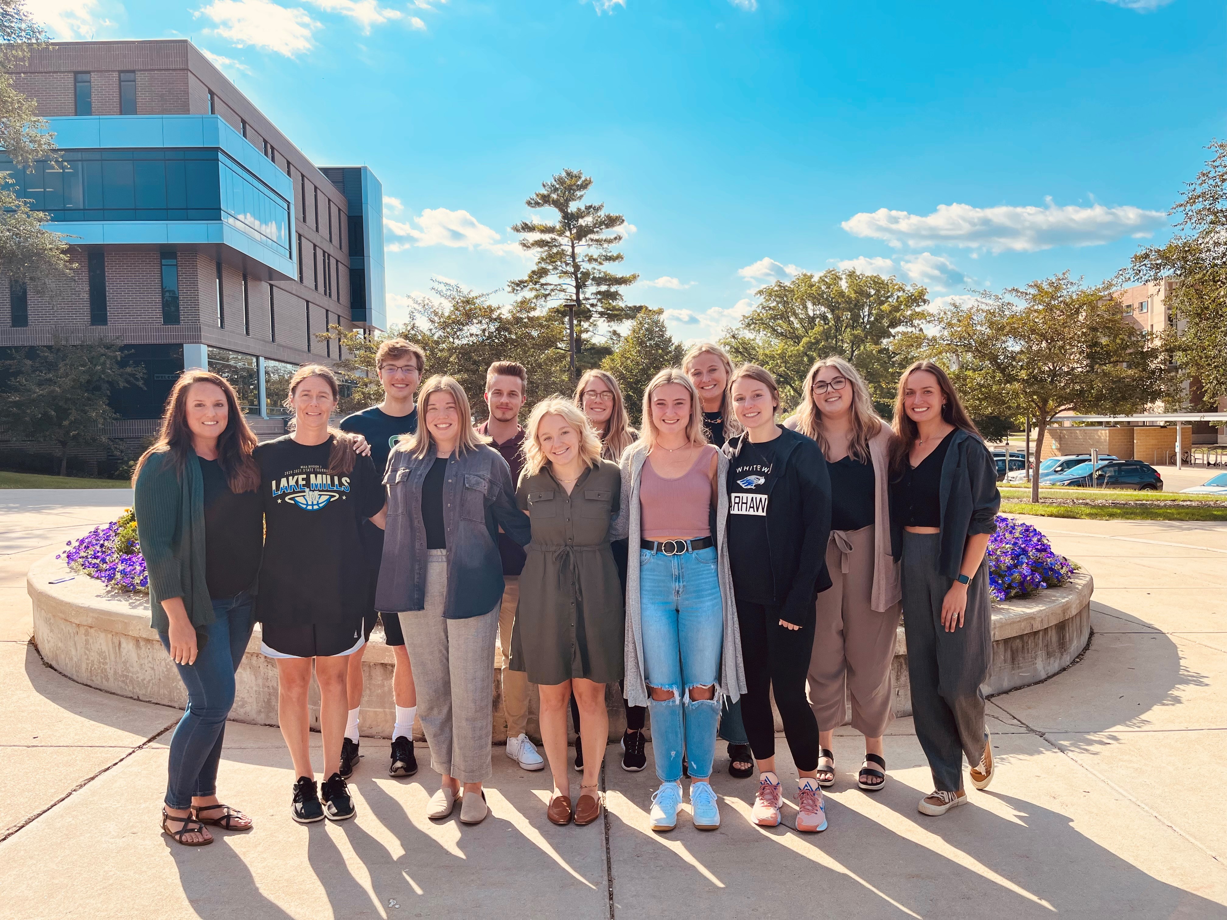 A group of people stand together in a row outdoors on campus.