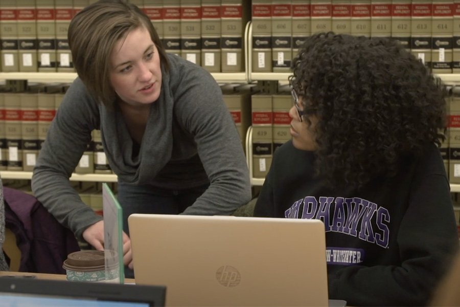 Two students discuss over books and a laptop.