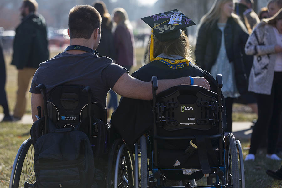 Two people outdoors in wheelchairs with arms around each other at graduation.